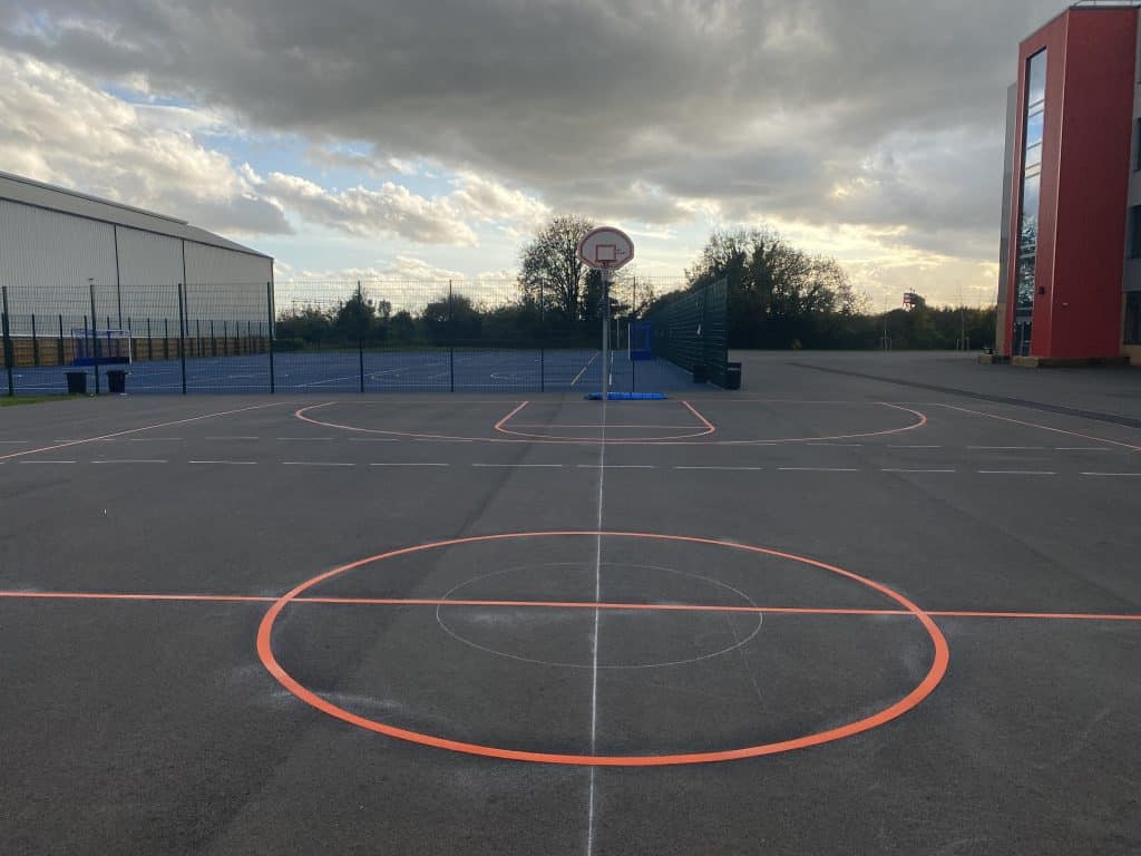 Orange basketball court markings and a hoop on grey tarmac, with a fenced blue MUGA and a red-clad building behind