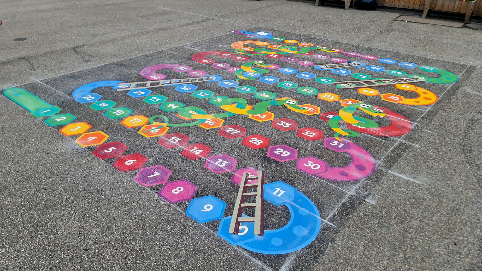 Snakes and ladders game with rainbow hexagon squares numbered 1 to 100 and smiling green snakes on grey playground tarmac