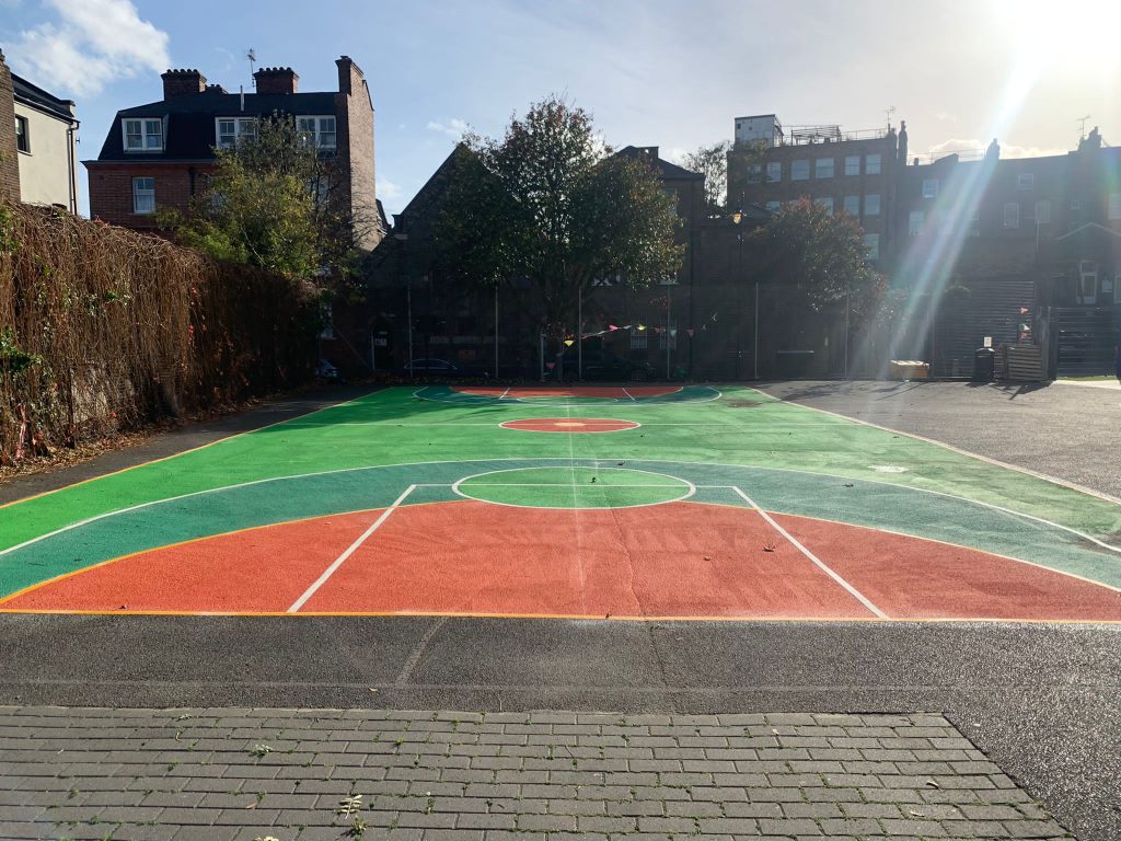 Basketball court with green, teal and orange surface coating and white lines, overlooked by brick houses and flats