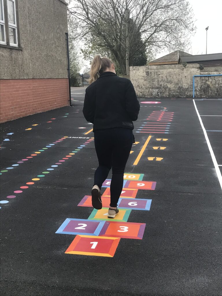 Woman in black hopping along a rainbow square hopscotch on new tarmac, with coloured spot trails and a sprint lane beyond