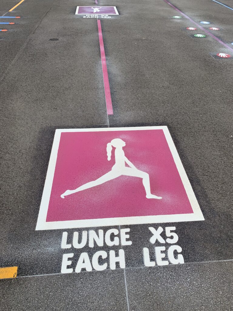 Pink fitness trail station with a white lunge figure and the words Lunge x5 each leg, and a hop station further along