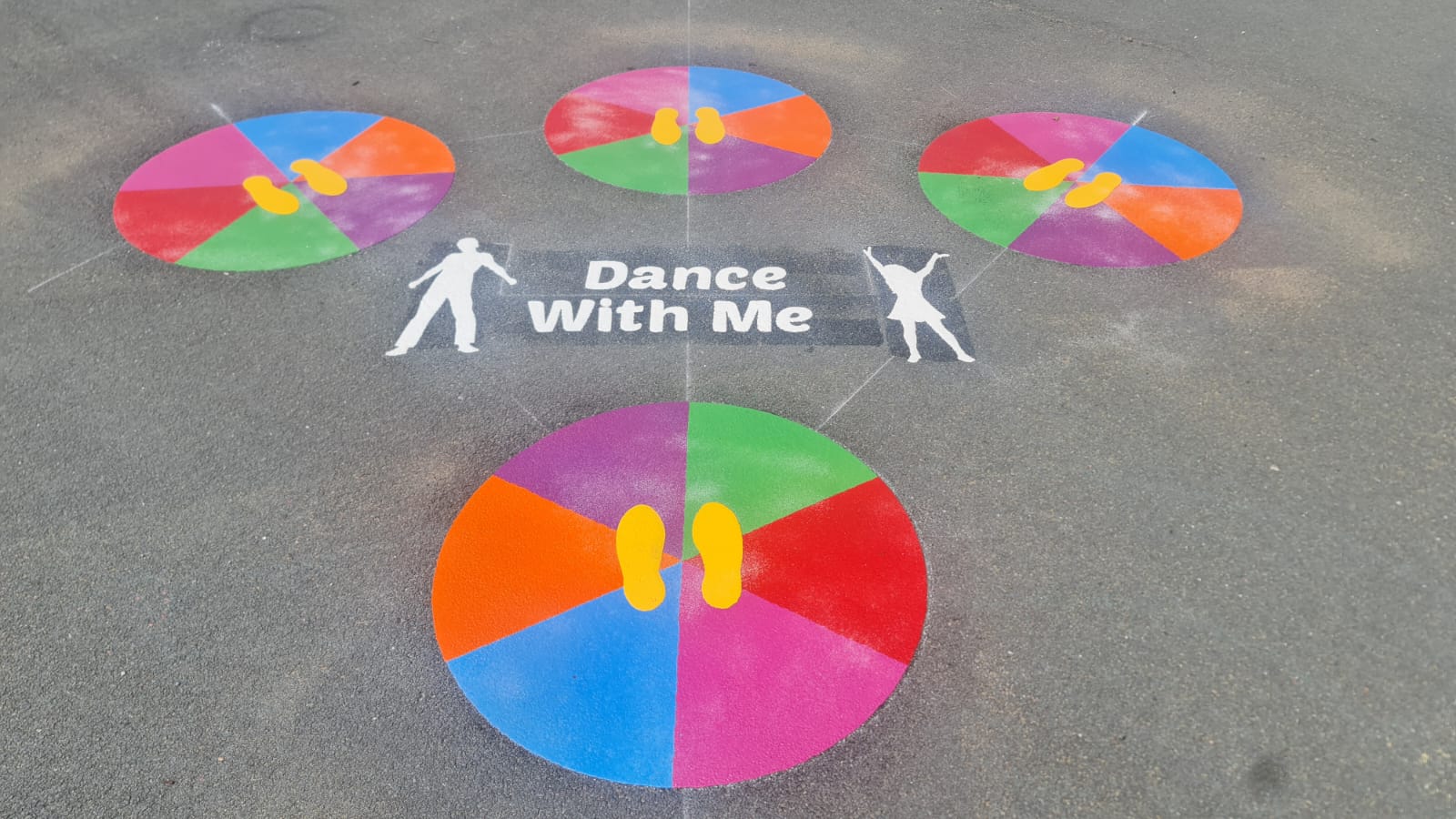 Dance With Me marking with four rainbow circles holding yellow footprints and two white dancing figures on grey tarmac